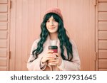 Small photo of Stylish young smiling hipster woman with color hair wearing trendy peach color coat and hat with reusable coffee cup on wooden background. Urban seasonal city street fashion. Color of the 2024 year.