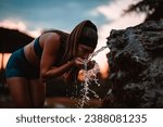 Small photo of An exhausted, fit girl drinking water from a fountain in the park after strenuous training. Her muscular built and athletic body shape showcase her active and healthy lifestyle.